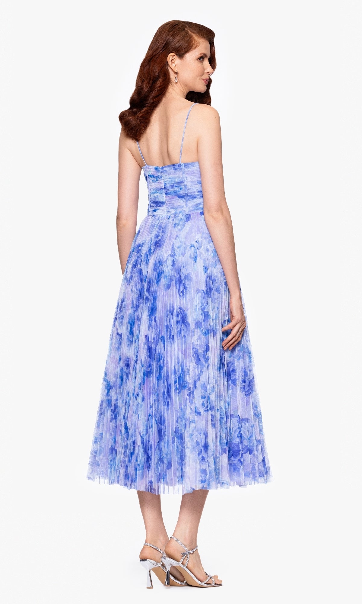 A woman with long wavy hair wears the Betsy & Adam A27053 midi wedding guest dress, which features a blue floral pattern, pleated A-line skirt, and strappy high-heeled sandals, standing with her back to the camera.